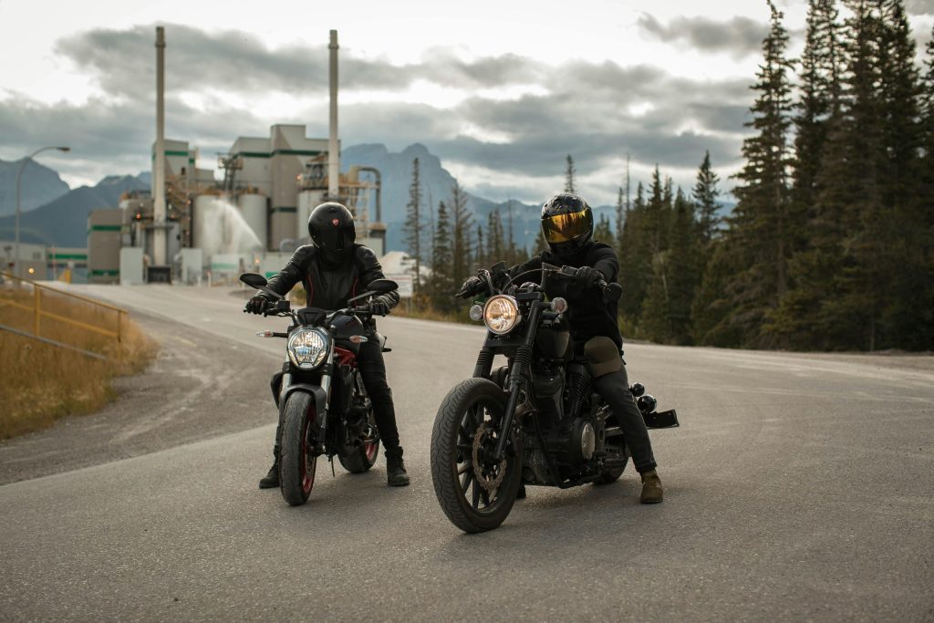 Two bikers in helmets on a road with forest and industrial backdrop riding motorcycles