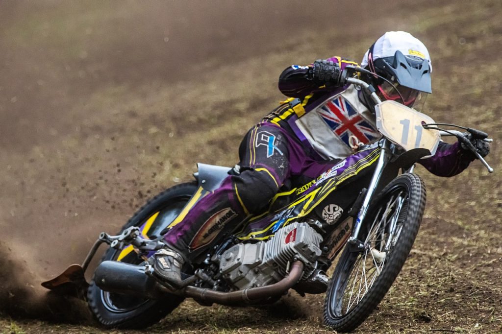 Motorcyclist racing on a dirt track wearing purple gear with Union Flag design, displaying dynamic motion and speed