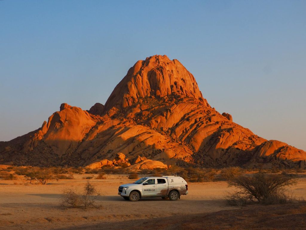 White truck parked in a desert landscape with a large red rock formation and blue sky in the background